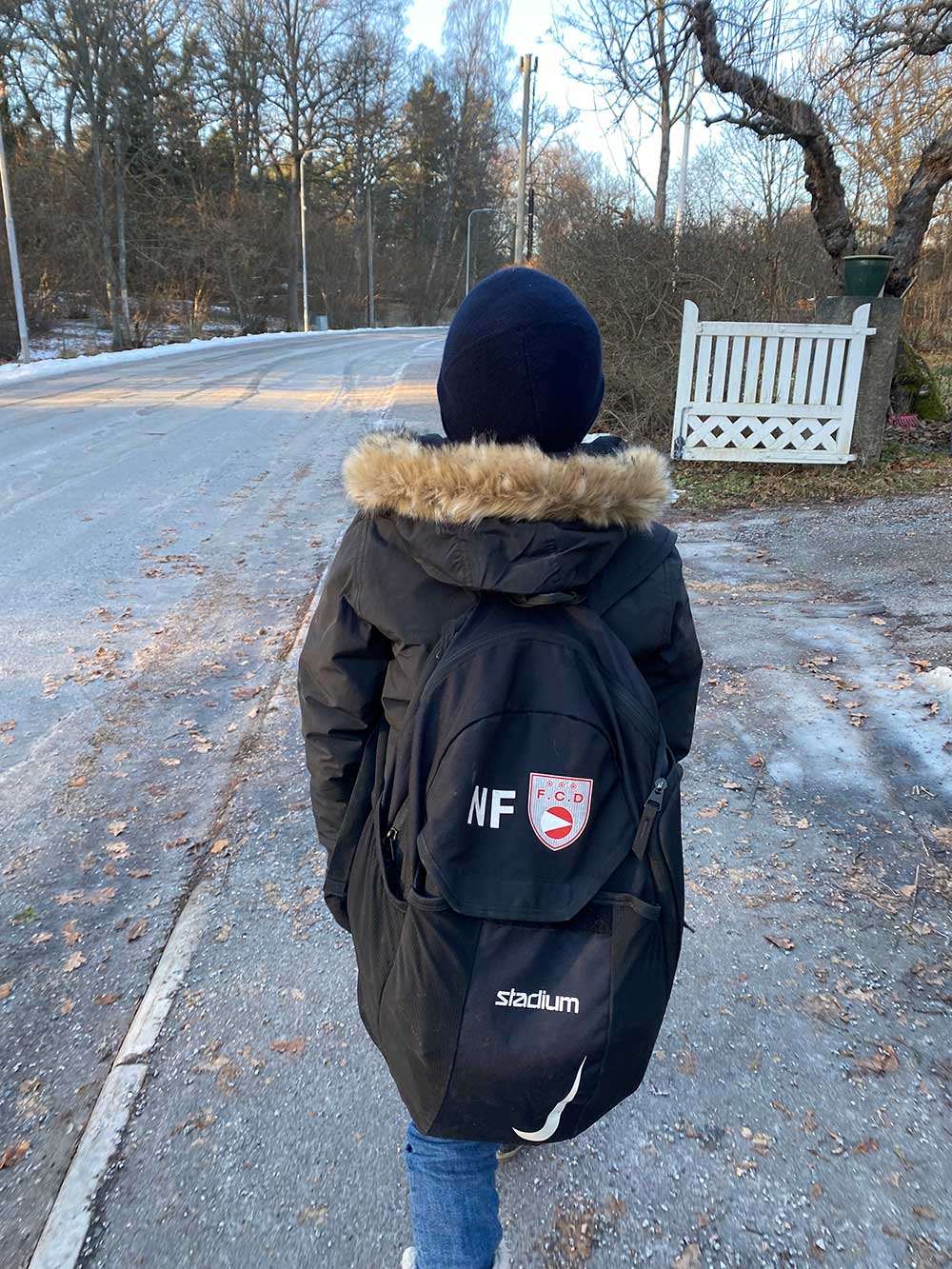 Ett barn i svart vinterjacka går ute på en gata med en svart ryggsäck och mössa